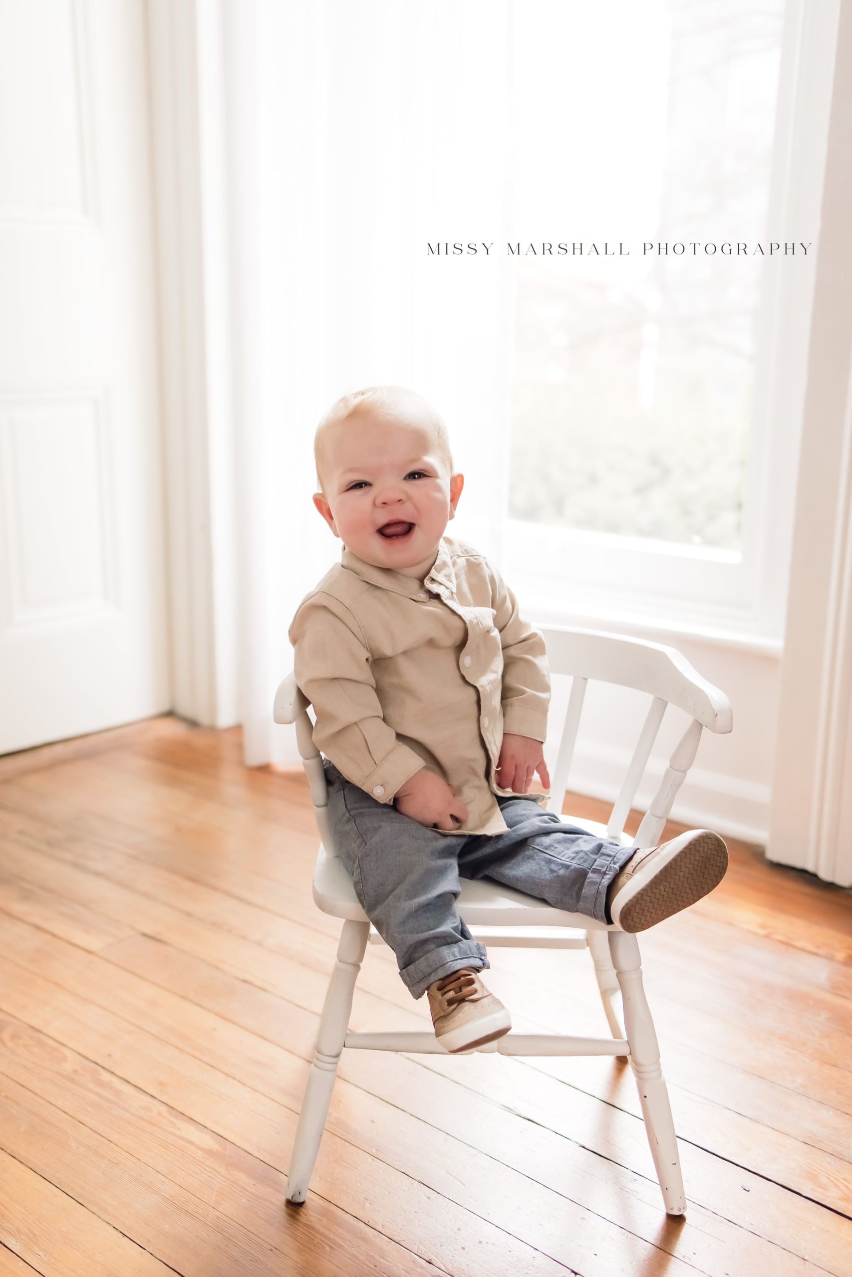 One year old baby smiling while sitting on a white chair in a bright studio in Louisville KY taken by Missy Marshall Photography