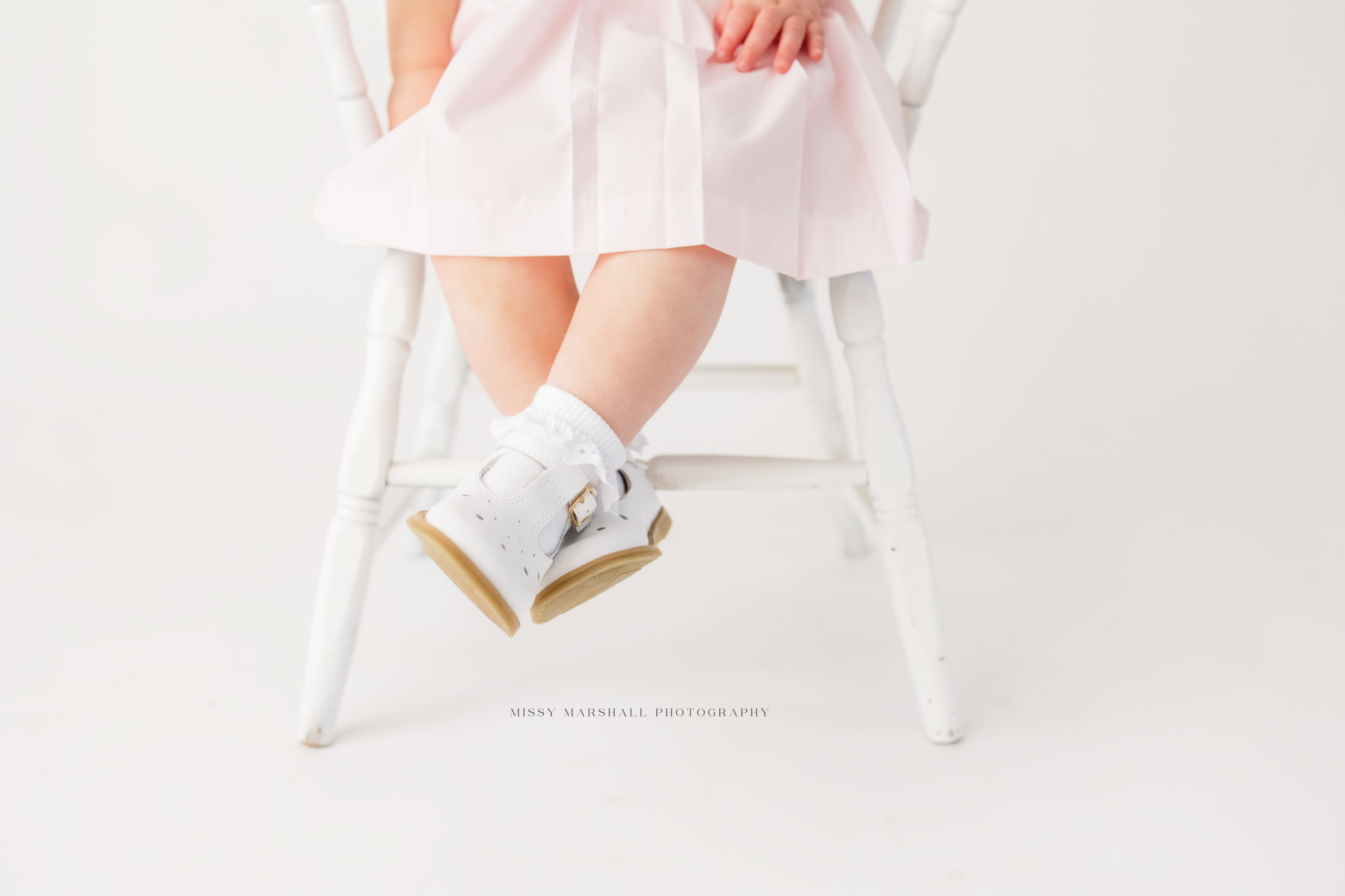 Baby sitting on a white chair with white shoes on her crossed feet taken by a baby photographer in Louisville