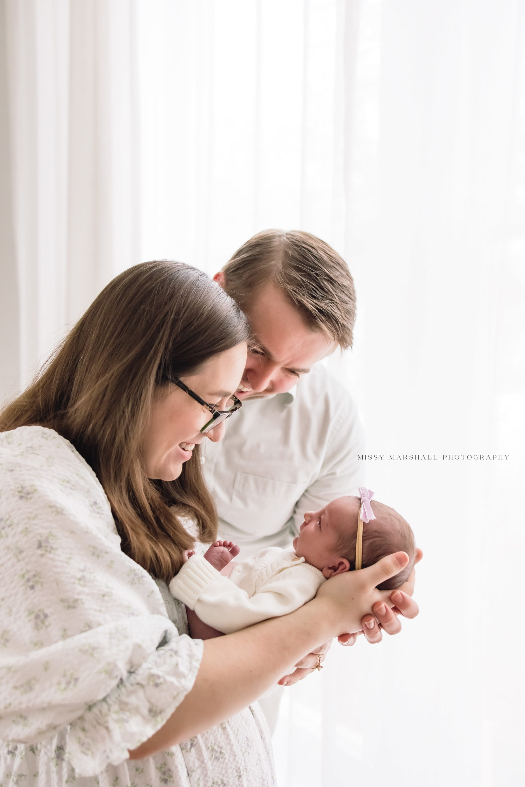 First time mom and dad holding their newborn baby girl and smiling lovingly in a bright studio in Louisville.