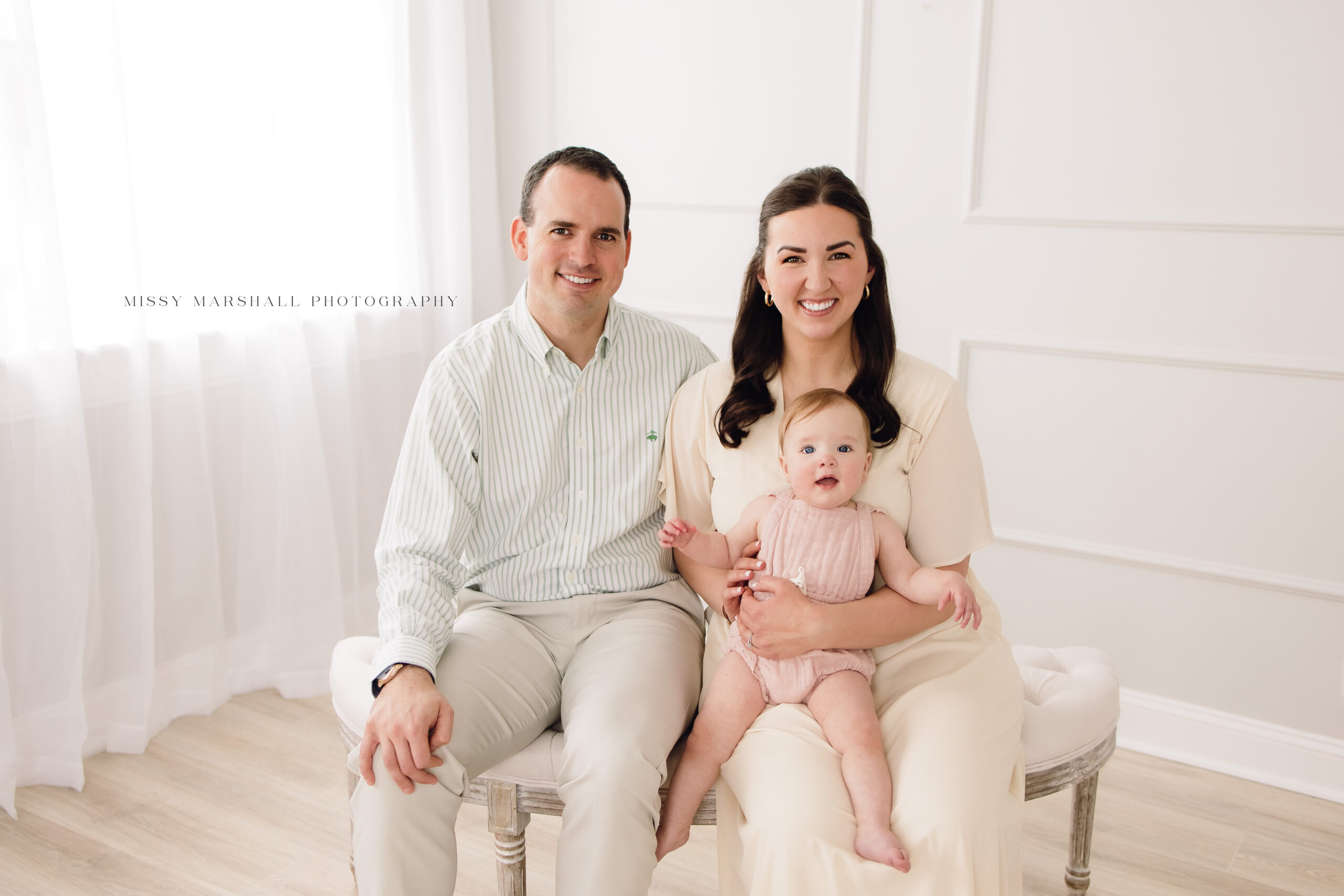 Mom, dad, and baby girl sitting on a neutral bench in front of a bright window during their session with a Louisville milestone photographer