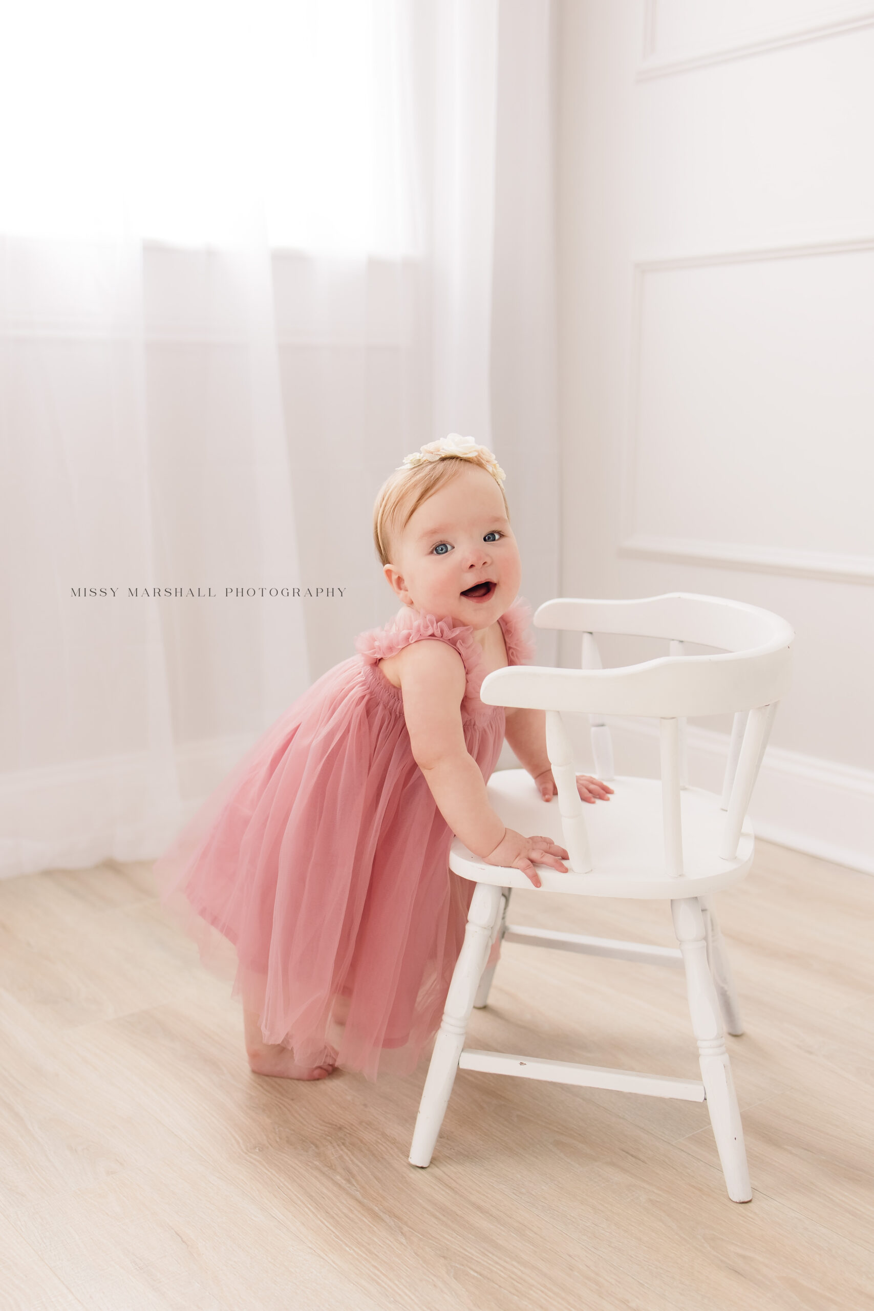 One year old baby girl wearing a pink tulle dress and standing up holding on to a white wooden chair in a beautiful bright studio
