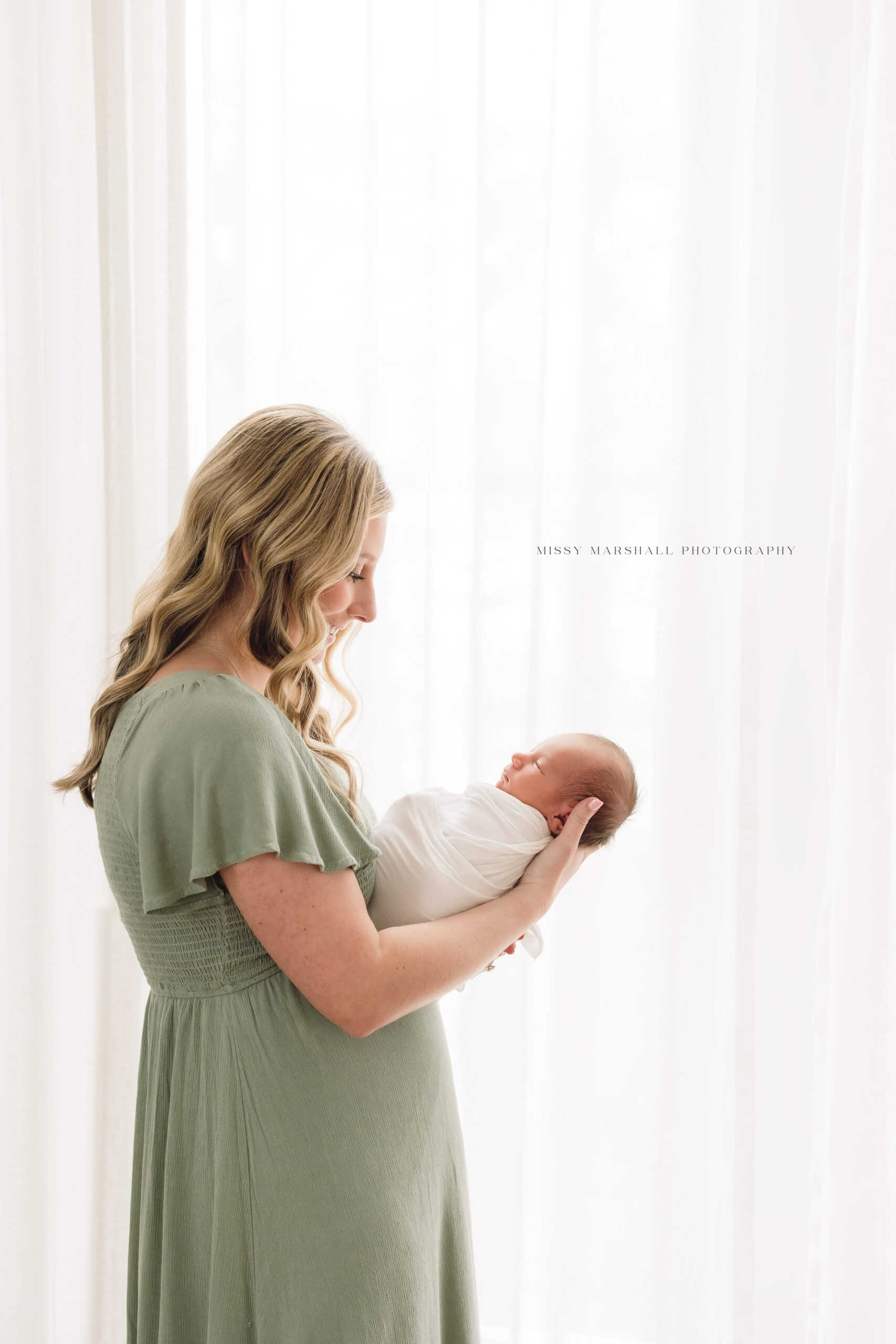 New mom smiling and holding her newborn baby in front of a window taken by a Louisville newborn photographer
