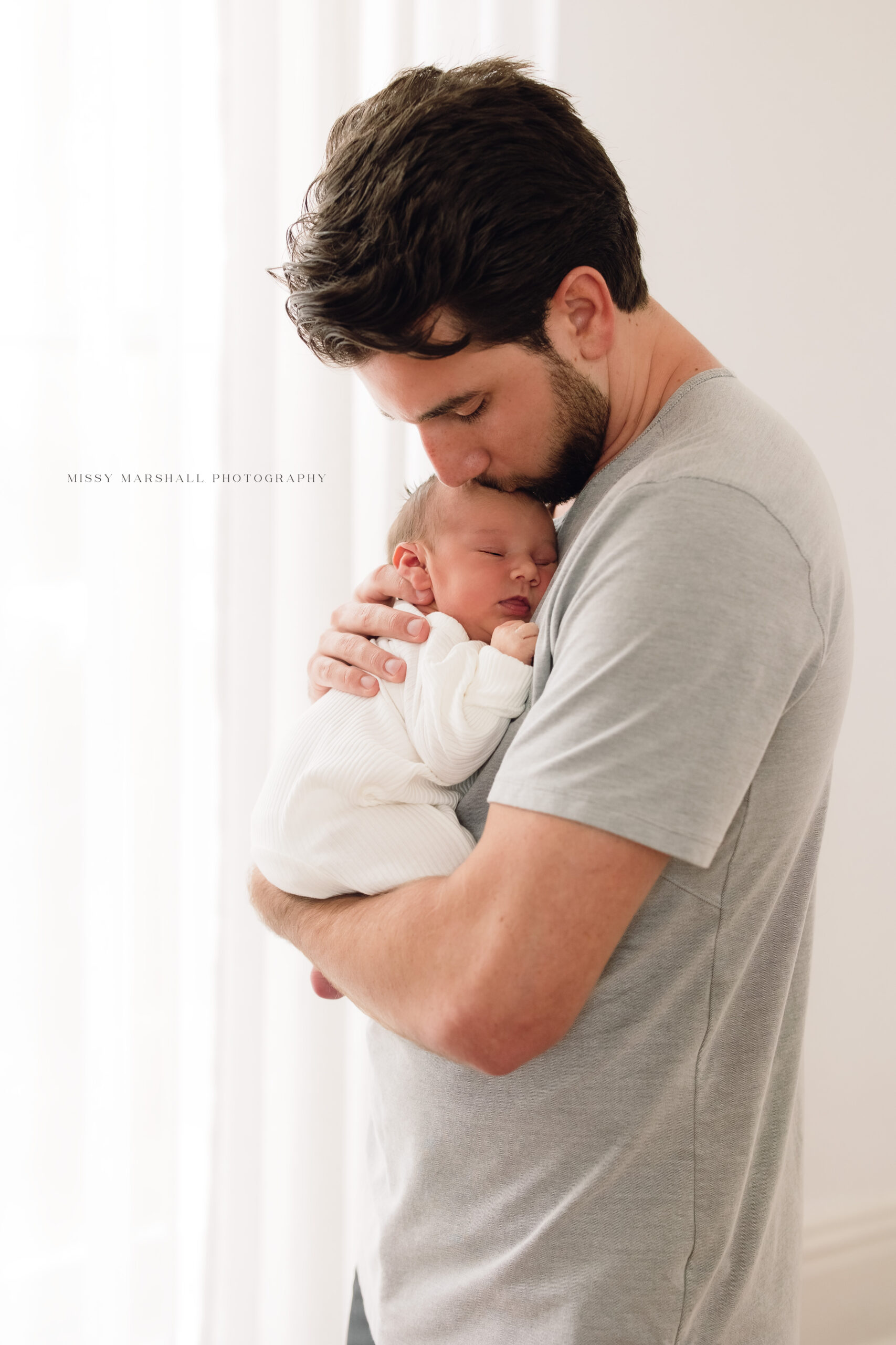 New dad kissing his newborn baby on his head in front of a window taken by a Louisville newborn photographer