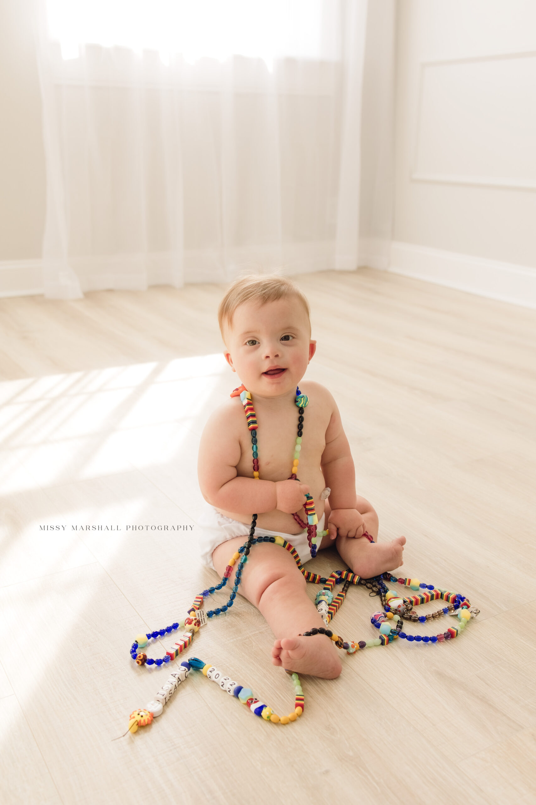 Baby boy dressed in a white diaper is sitting on the floor covered in a strand of multi-colored beads