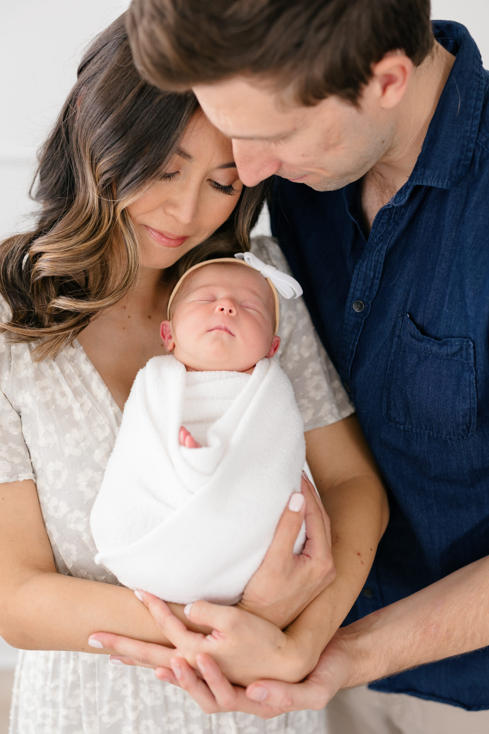New mom and dad snuggling their sleeping baby girl who is wrapped in a white swaddle