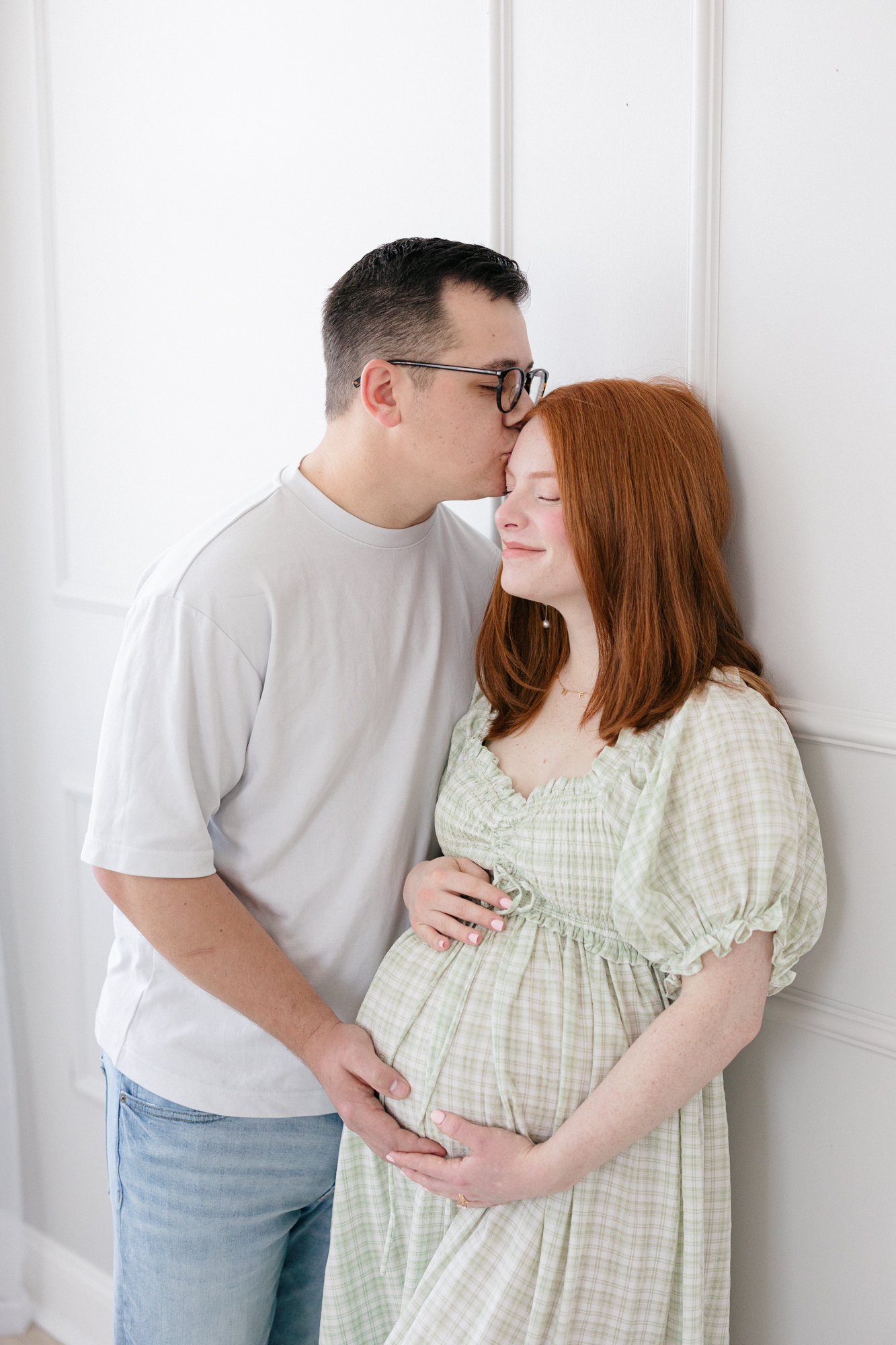 Expecting parents embracing against a wall in a bright Louisville KY photography studio